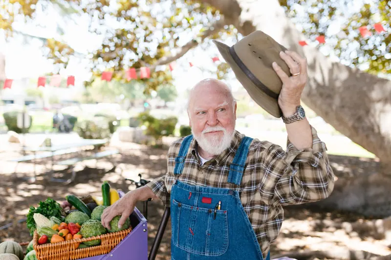 A grandfather in overalls waving his hat beside a cart of garden vegetables