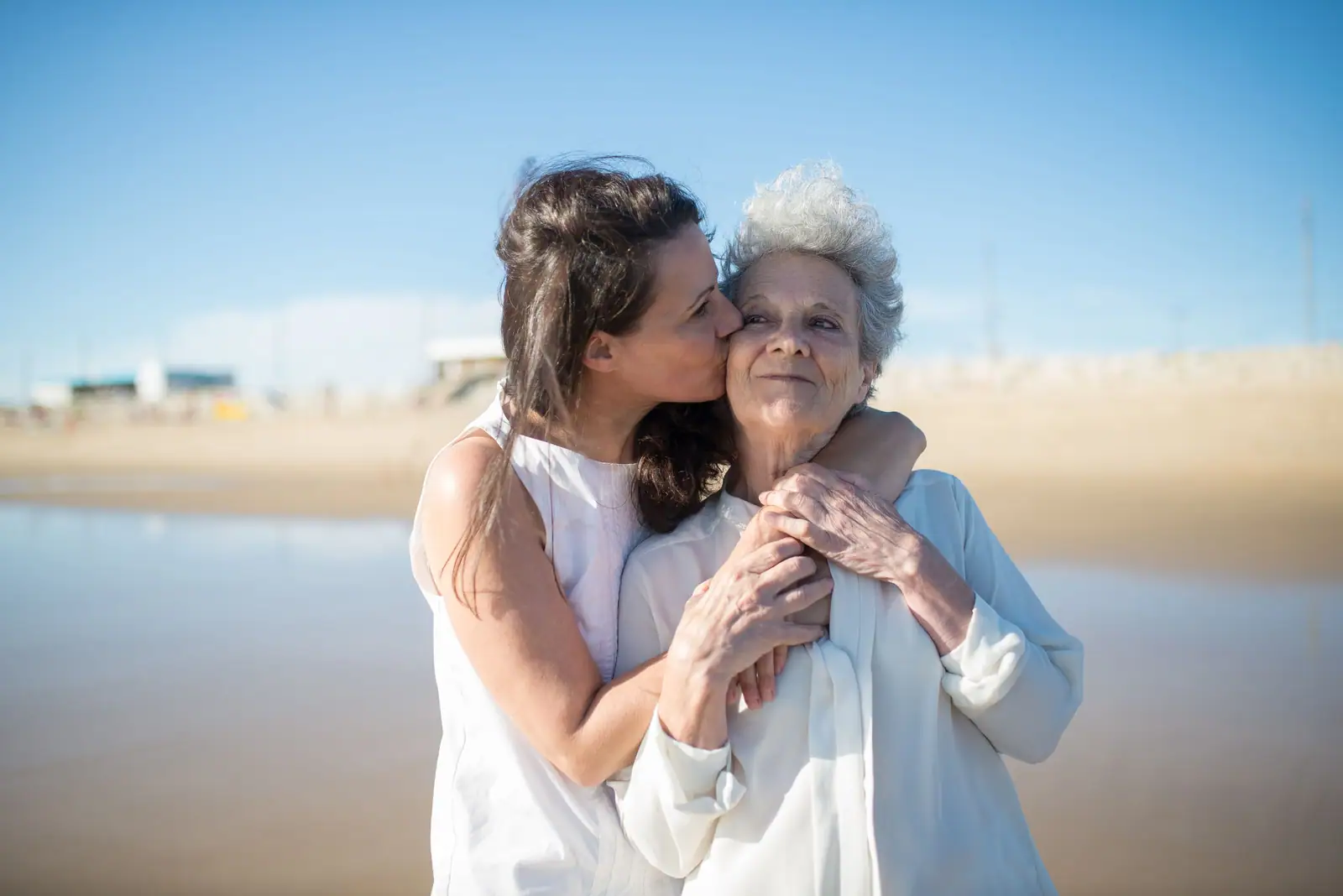 A daughter kissing her mother's cheek on a sunny shoreline