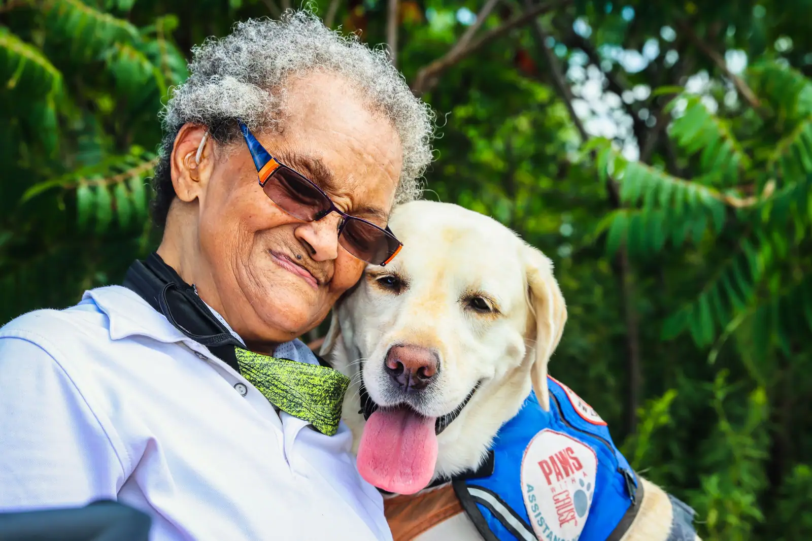 A smiling resident in sunglasses hugging a yellow Lab in a blue therapy vest, cheek to cheek in the garden