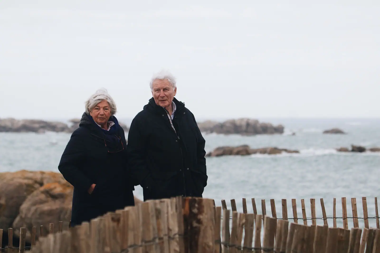 Elderly couple stands together near the ocean, enjoying a peaceful outing.