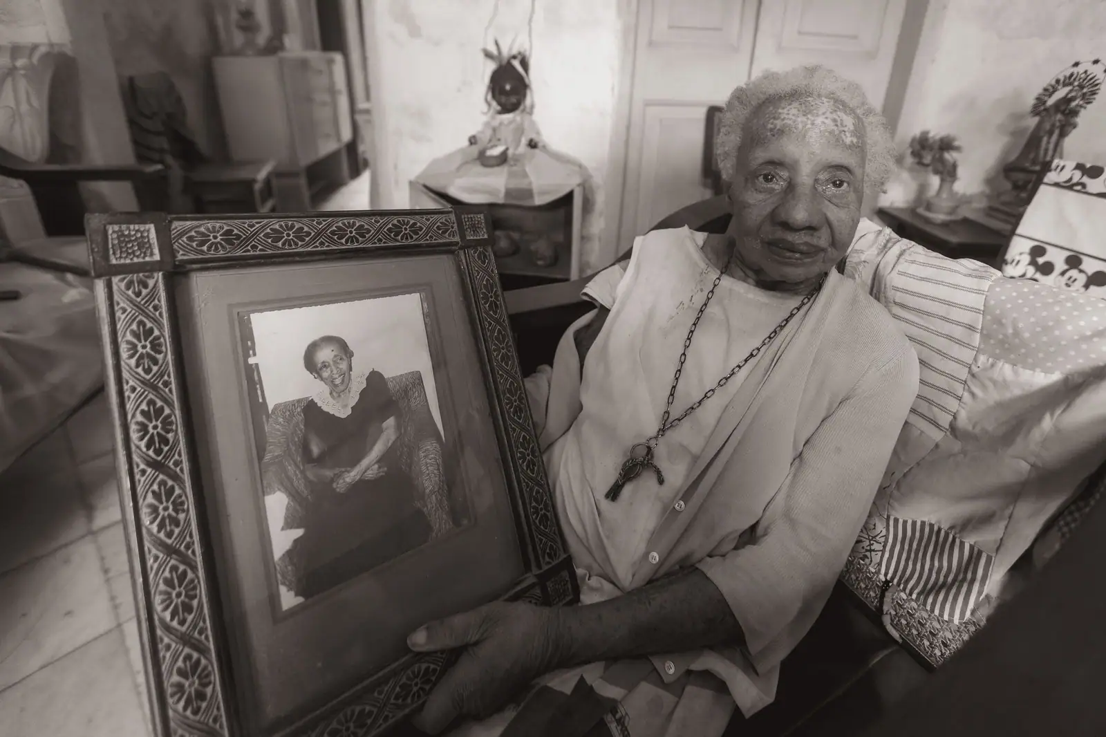 An elderly woman holds a framed photograph of herself in a cozy living room.