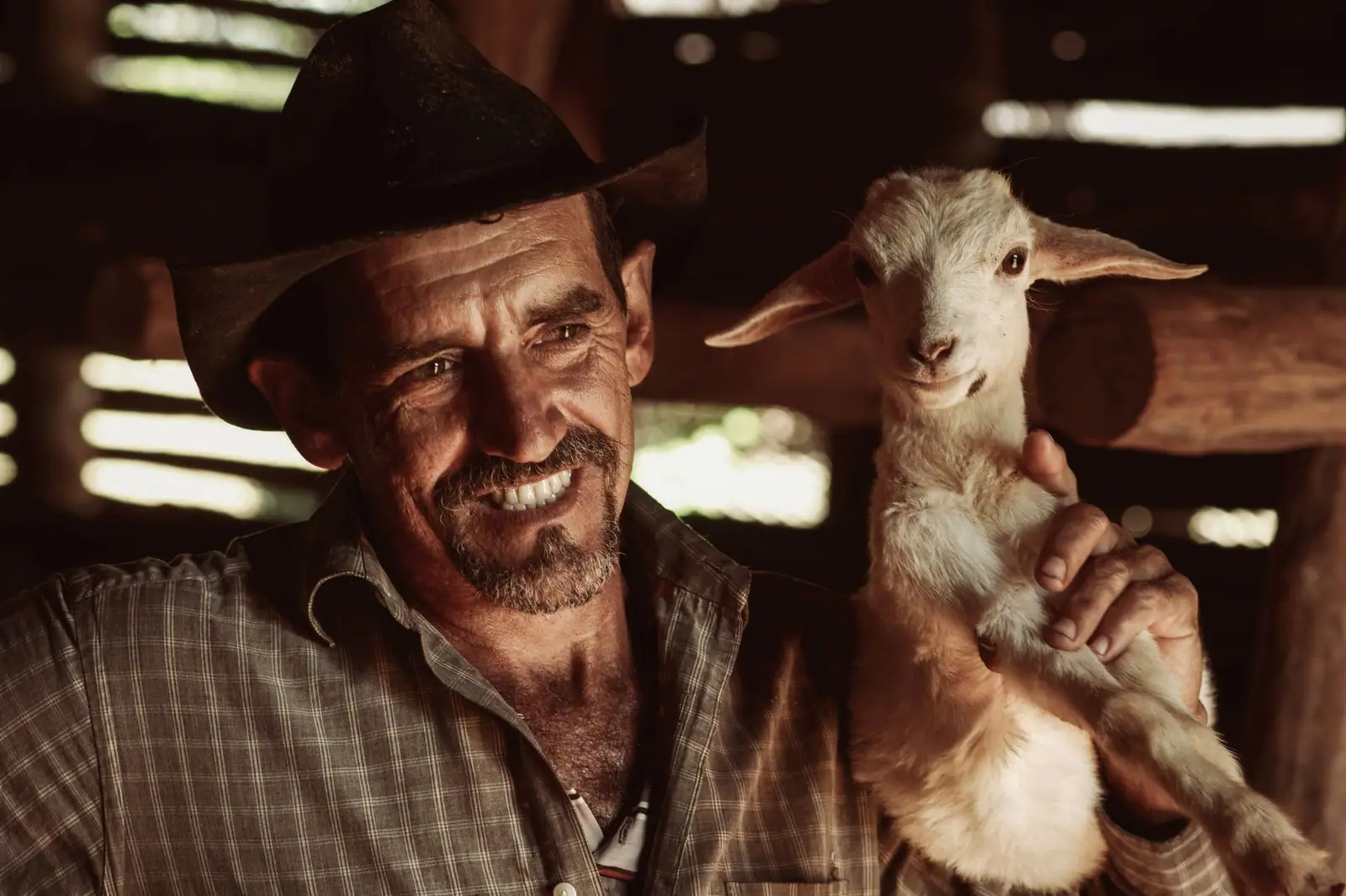 Smiling farmer holding a young goat inside a rustic barn. The warm lighting adds to the intimate farmhouse setting.