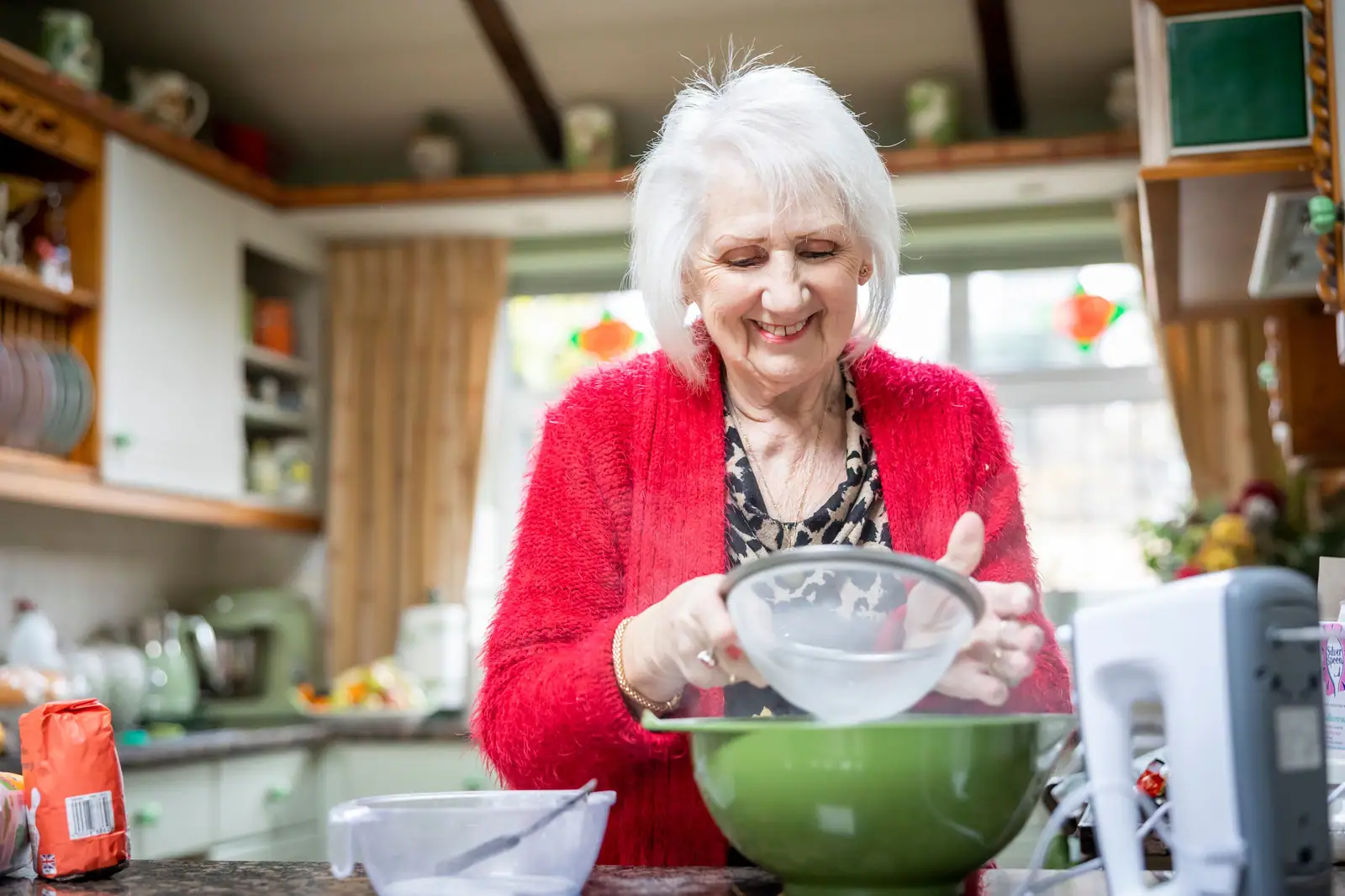 A smiling resident in a red cardigan sifting flour into a mixing bowl in a bright kitchen