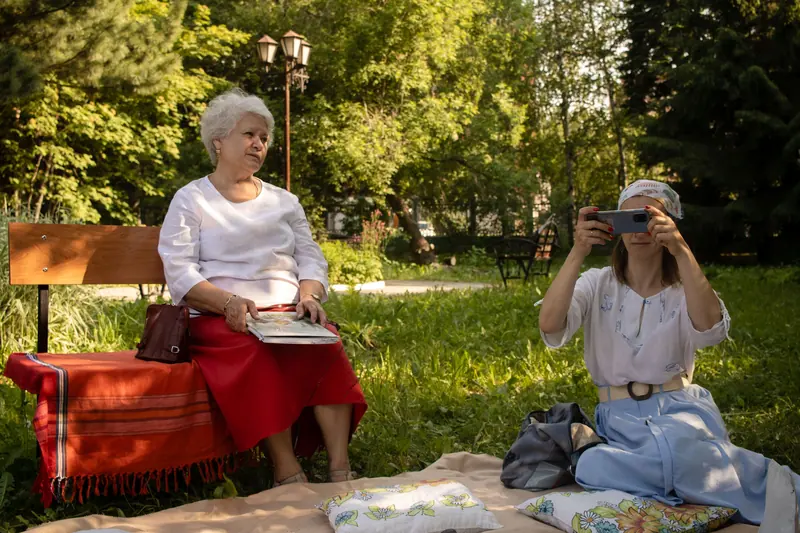 A resident reading on a picnic blanket while her granddaughter takes pictures