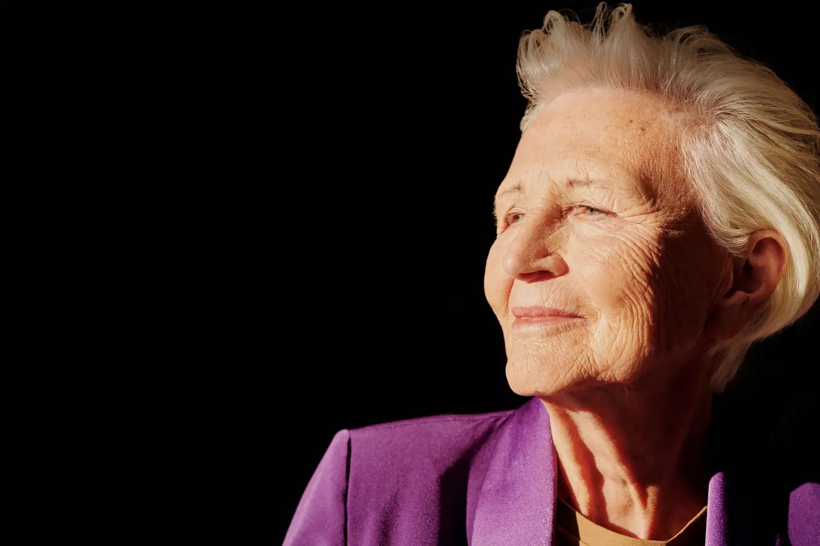 A resident with silver hair and a violet jacket, eyes half closed and smiling in a shaft of evening light against a black wall