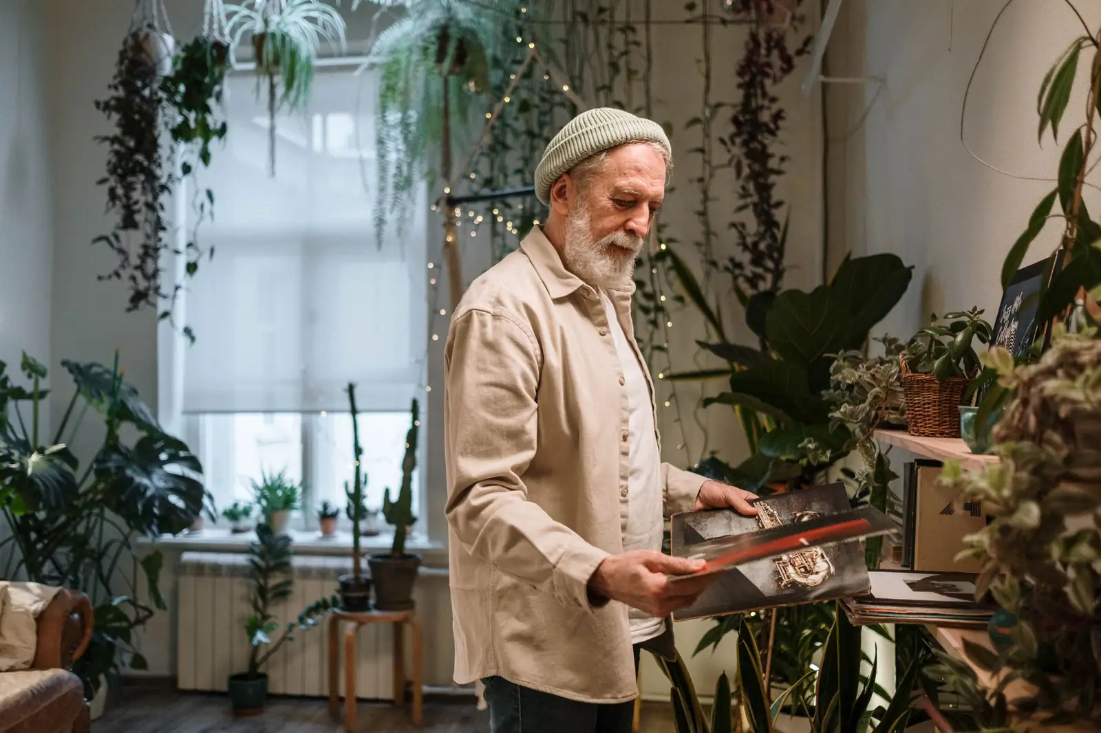 Senior man with grey hair in indoor garden, surrounded by plants, holding vinyl records, enjoying a vintage moment.