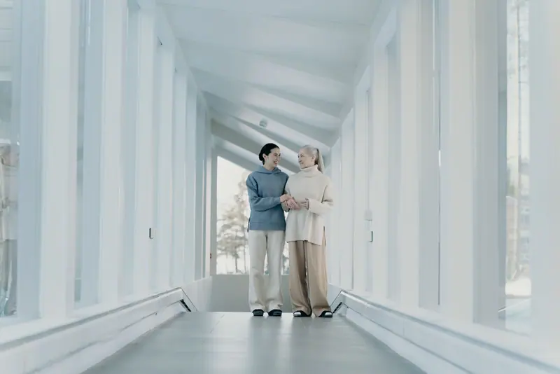 A caregiver and a resident walking arm in arm down the bright hallway