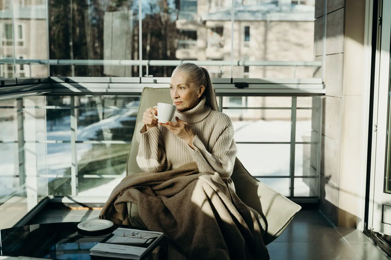 A resident in a cream sweater holding a cup of coffee by a sunny window, a blanket over her knees