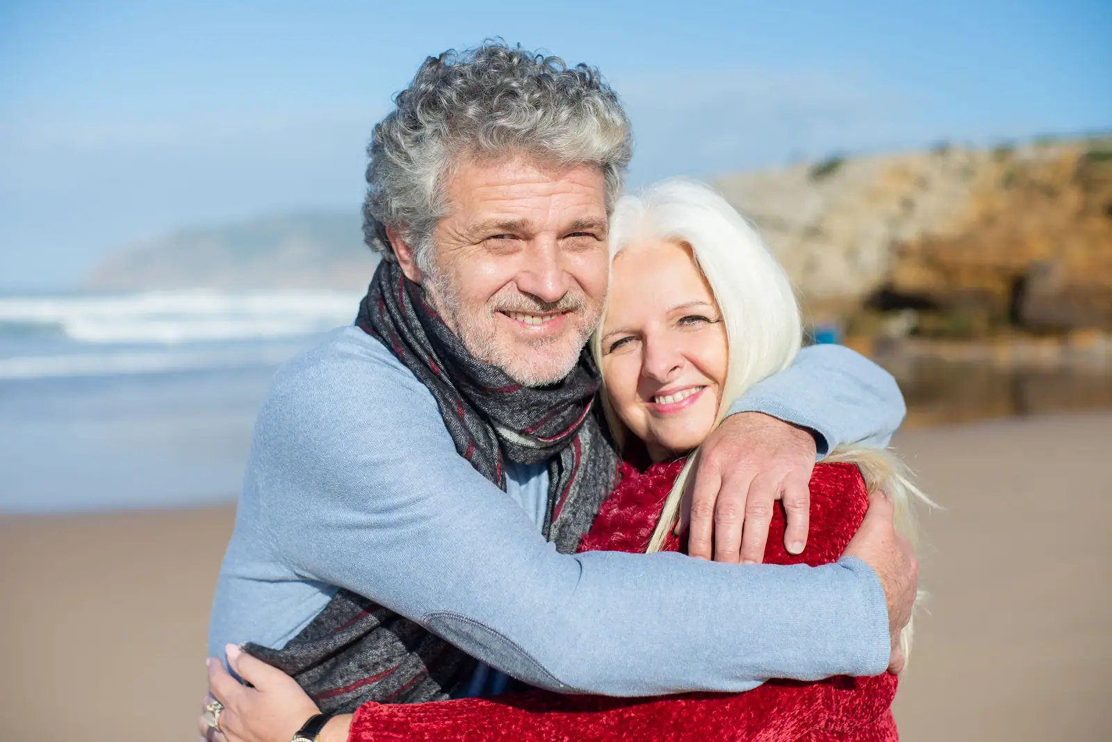 A silver-haired husband and his wife hugging and smiling on the beach on a breezy blue day