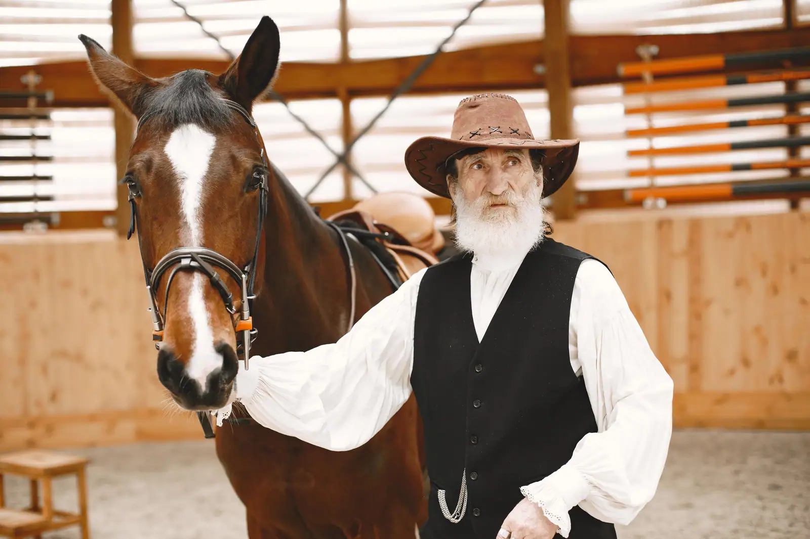 Elderly man in cowboy hat beside a brown horse, standing in a rustic barn.