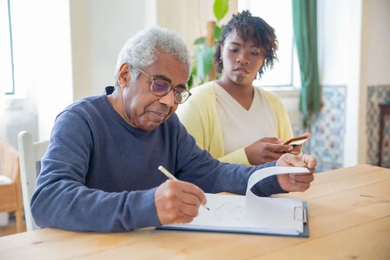 A resident working through a page of notes at the dining table while a caregiver looks on