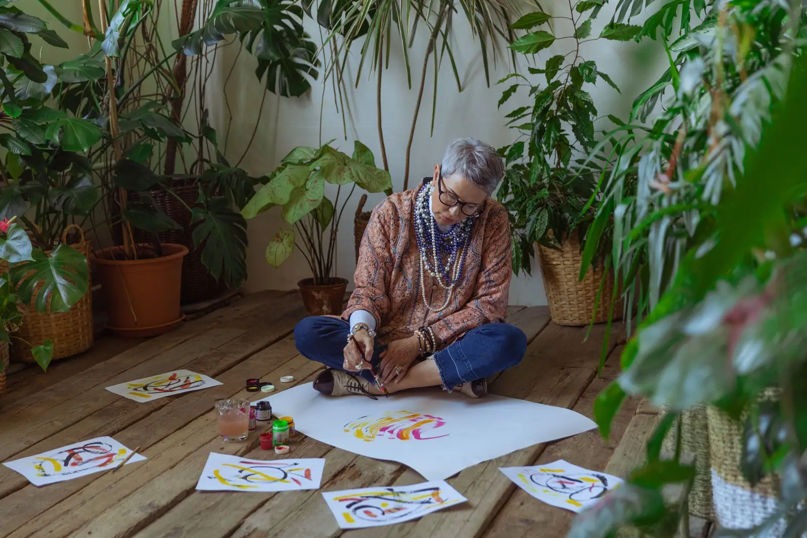 A resident sitting cross-legged on the studio floor among potted plants, painting bright brushstrokes on large sheets of paper
