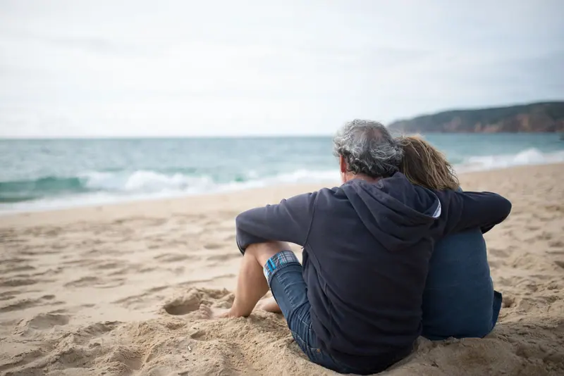 A couple sitting close together on the sand, looking out at the waves
