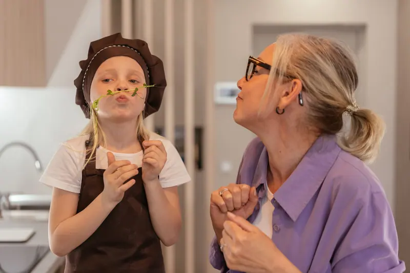 A caregiver and a girl making mustaches with pasta at snack time