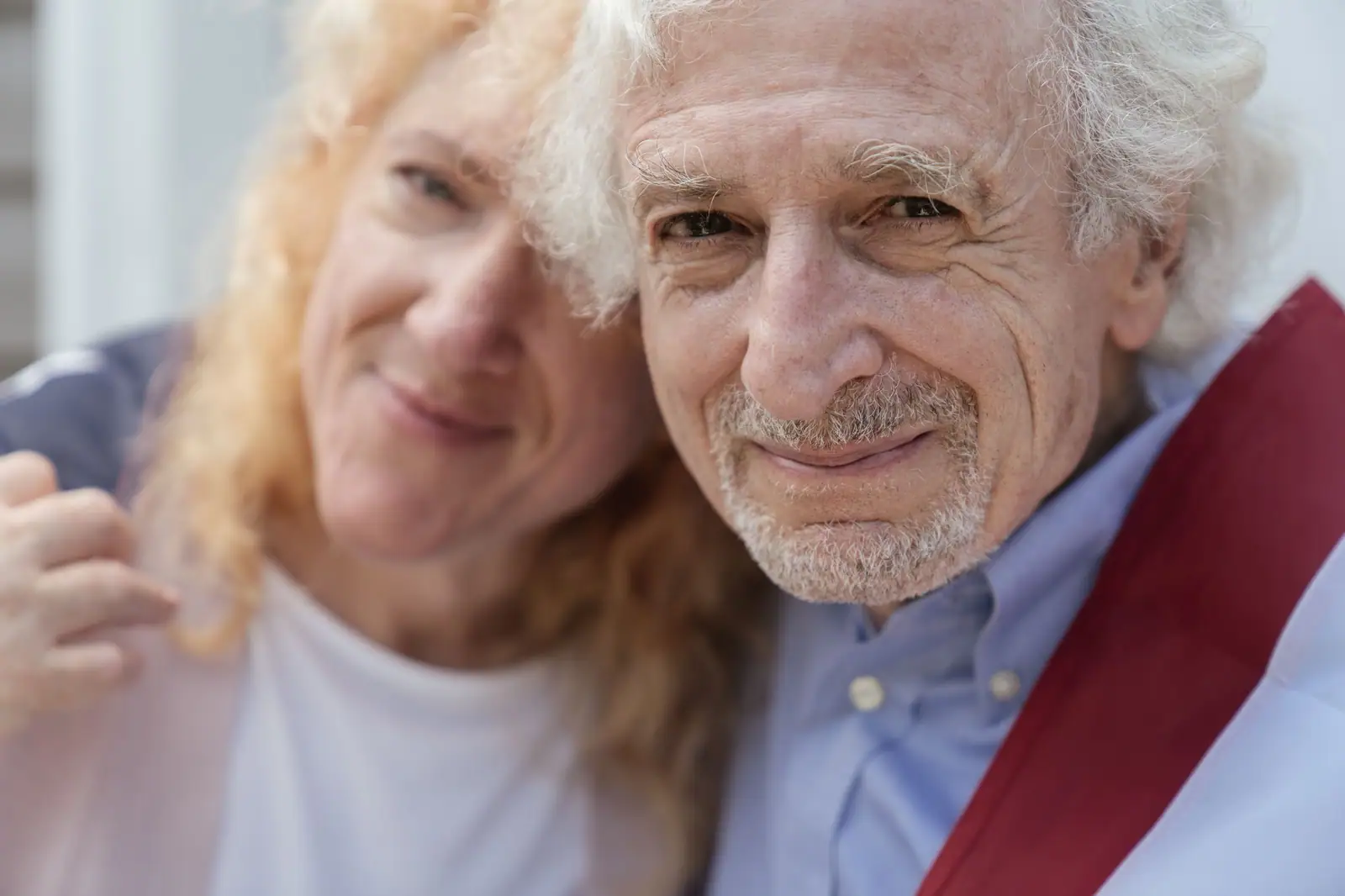 A veteran with a white beard and a folded flag across his shoulder, cheek to cheek with his smiling wife