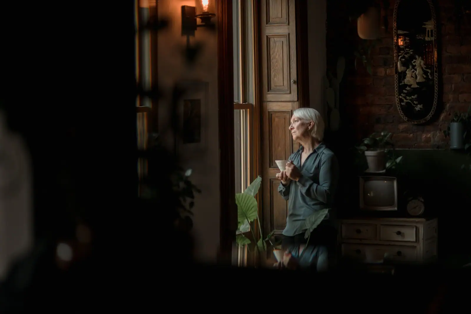 A resident in a gray cardigan standing by a tall window in a dim, lamplit room, a cup in her hand