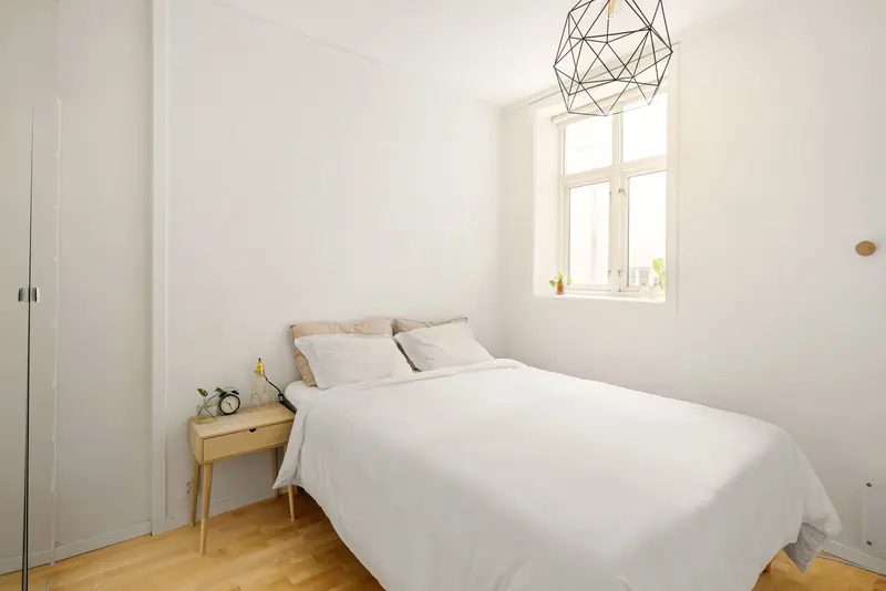 Bright minimalist bedroom featuring a white bed, wooden floor, and geometric pendant light.