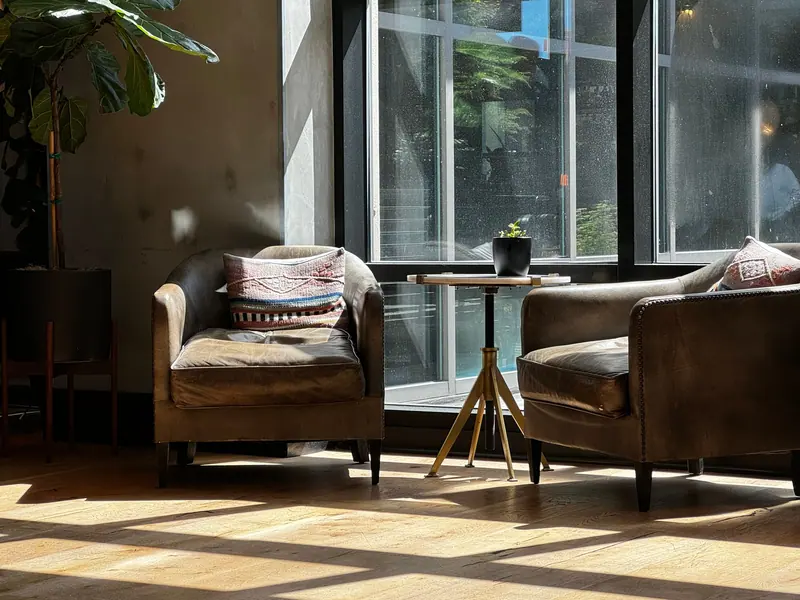 A cozy living room featuring two armchairs, a small table, and sunlight streaming through large windows.