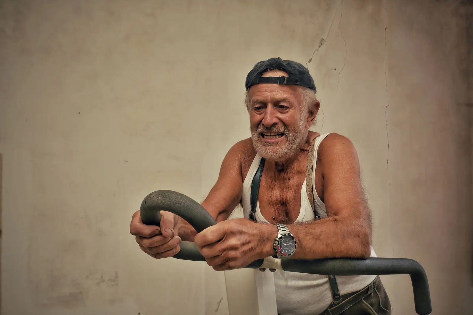 A smiling older man in a ball cap working out on the stationary bike in a darkened gym