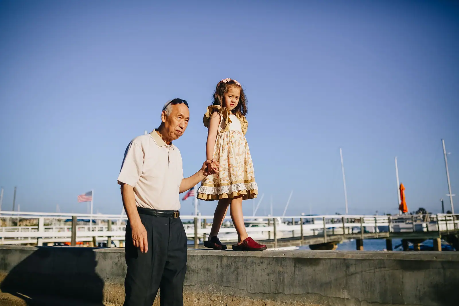 A grandfather holding his granddaughter's hand as she walks along a harbor wall on a sunny day