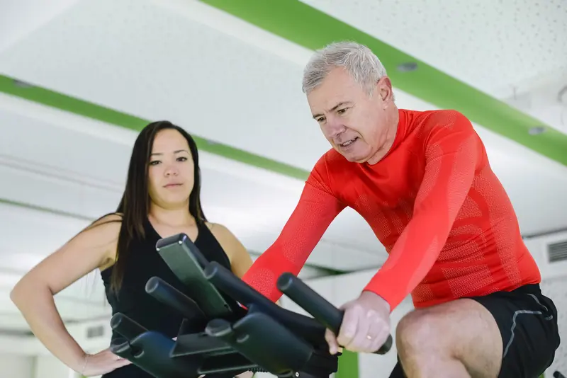 A guest on the recumbent bike with his therapist beside him
