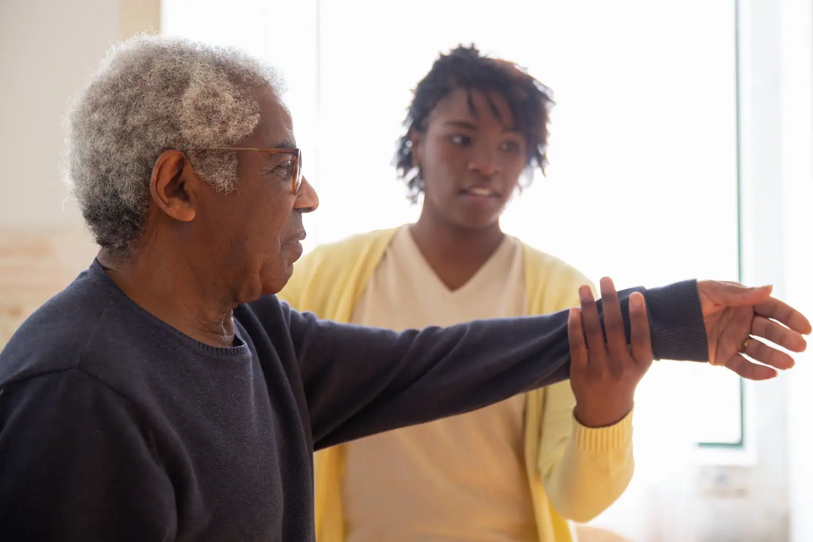 A therapist in a yellow cardigan guiding a resident through arm stretches by a bright window