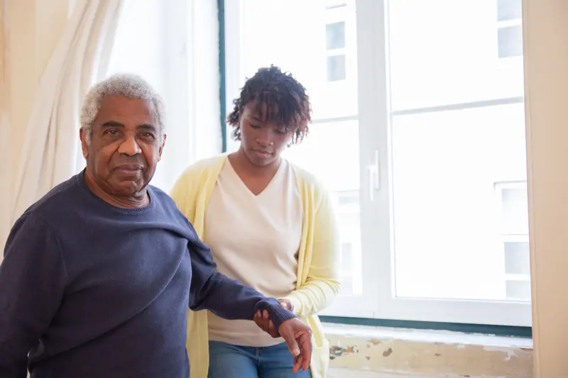 A caregiver steadying an older man as he stands from his chair