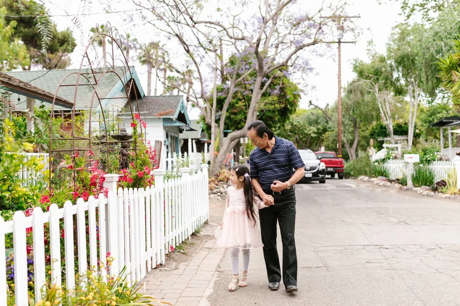 A senior man and young girl walking hand in hand on a suburban street lined with white fences.