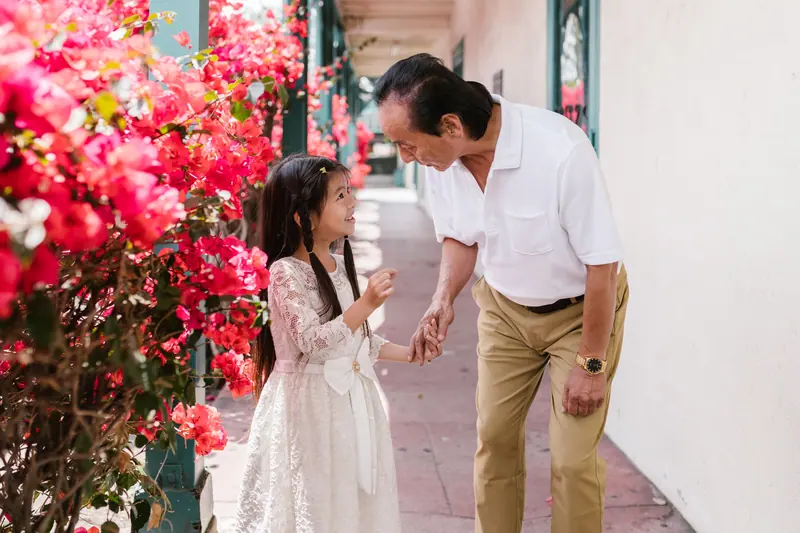 A grandfather bending down to talk with his granddaughter beside a flowering wall