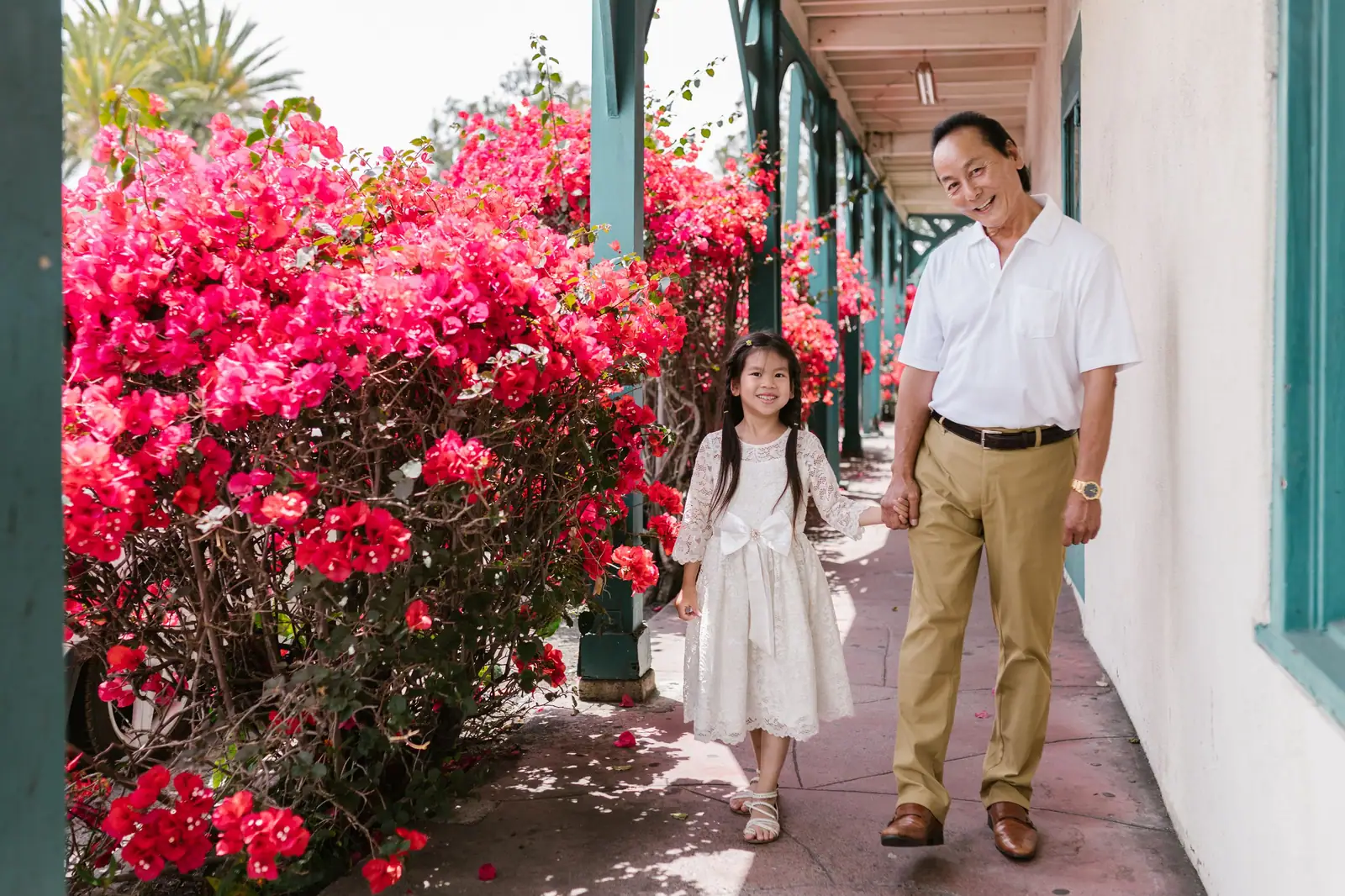 A grandfather walking hand in hand with his granddaughter past bright pink bougainvillea