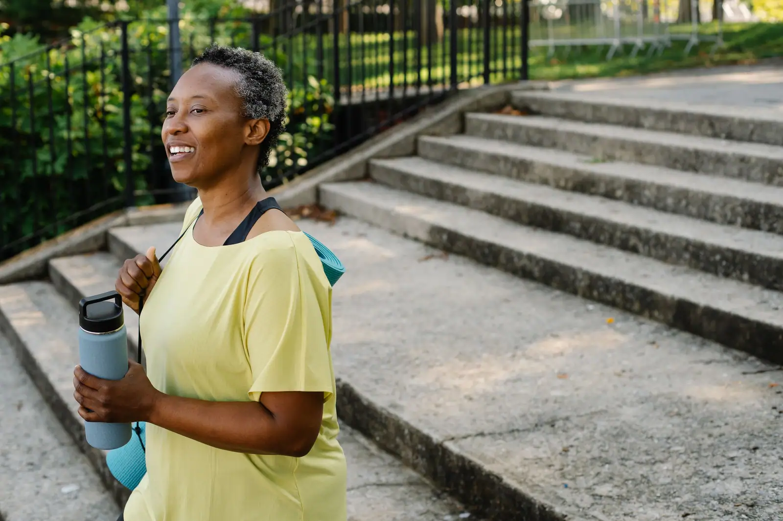 A smiling woman in a yellow T-shirt resting on park steps with her water bottle after a morning walk