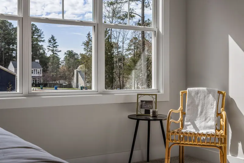 A cozy bedroom interior with a chair and a view of suburban homes through a large window.