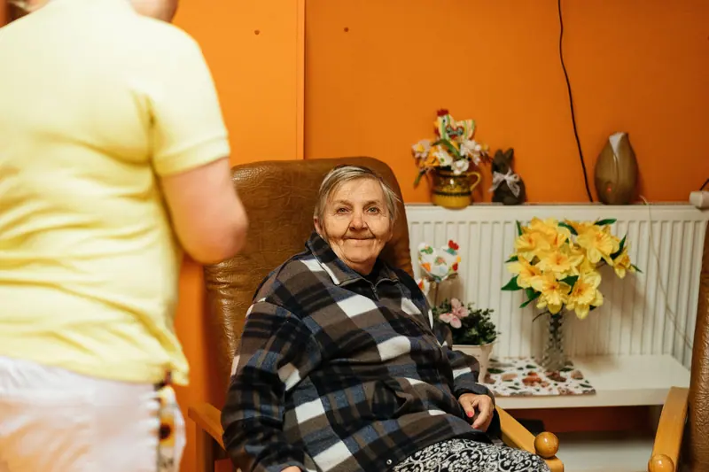 A smiling resident in a plaid jacket in her armchair, with a caregiver nearby