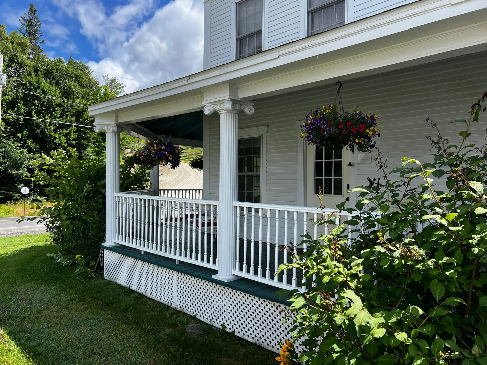A white farmhouse porch with hanging flower baskets and green lawn on a summer day