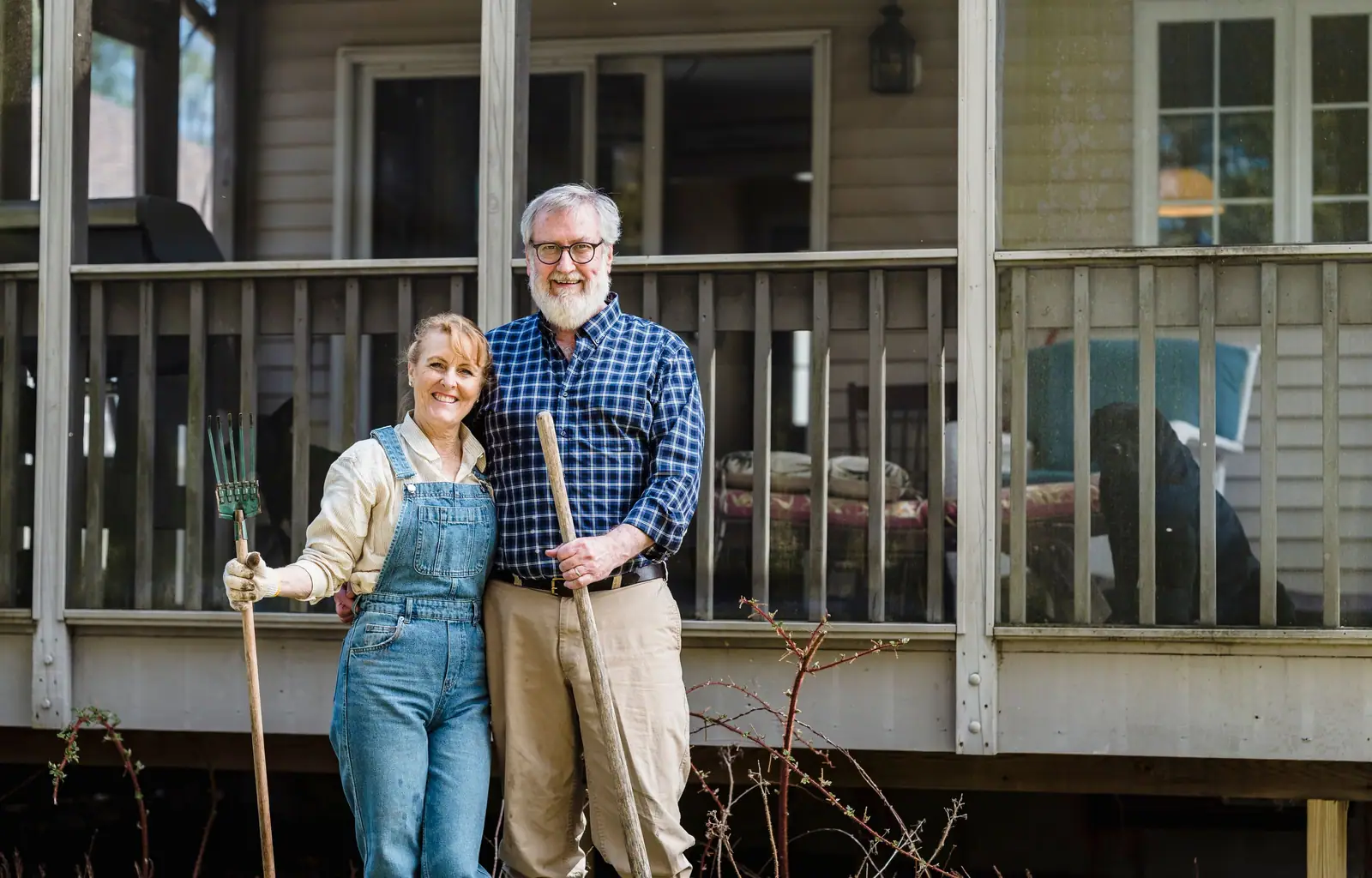 Lupe and Ray in front of the porch with a rake and hoe, ready for spring planting
