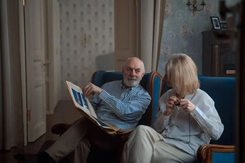A couple in the parlor, one knitting and one reading the paper