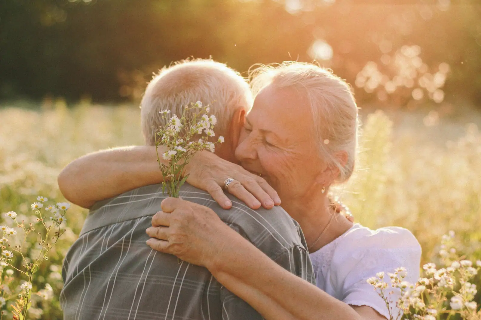 An older couple holding each other close in a sunlit meadow, she holding a handful of wildflowers