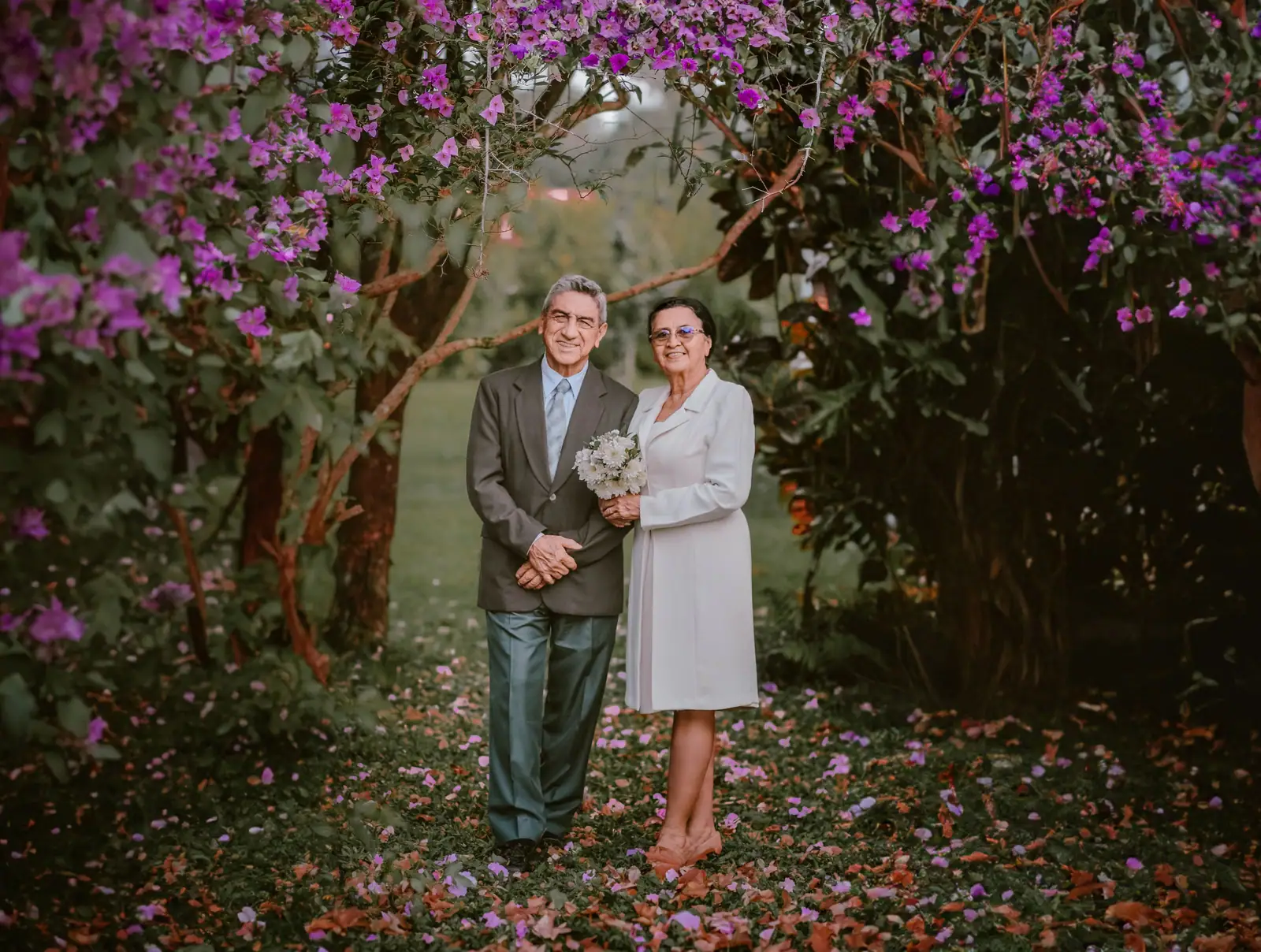 A smiling older couple standing arm in arm beneath a tree in full purple bloom