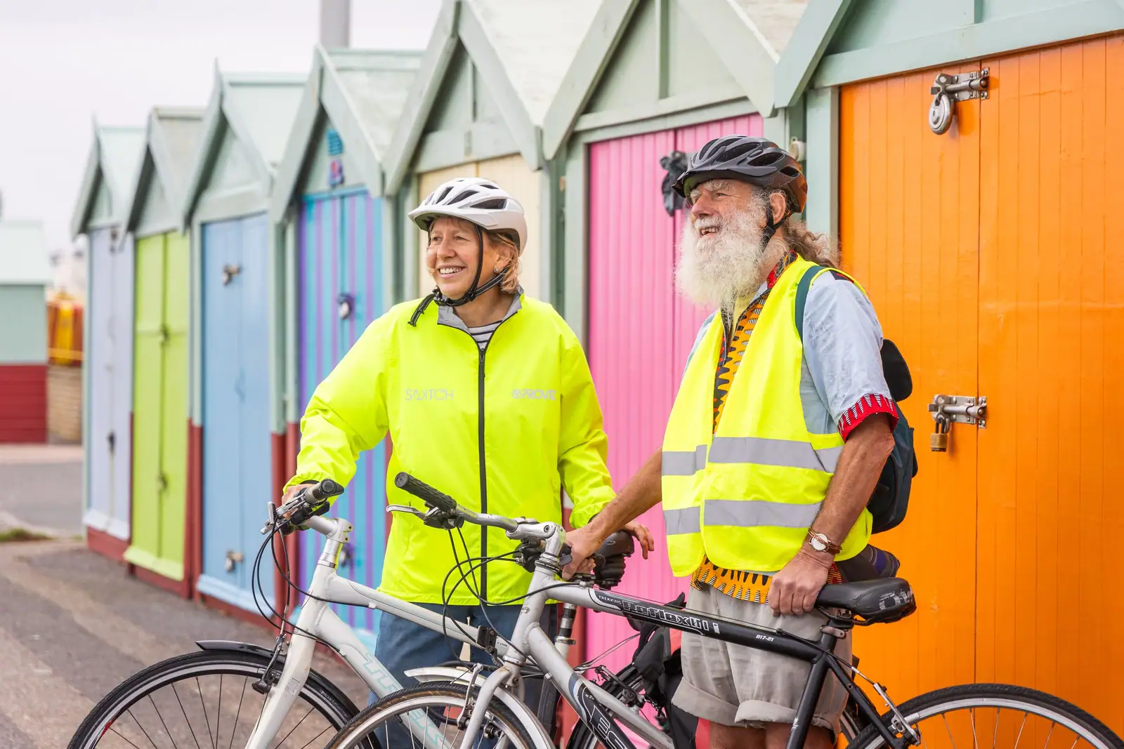 Senior couple enjoys cycling near vibrant coastal beach huts on a sunny day.