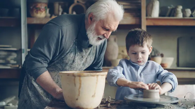 A resident teaching his grandson to center clay on the wheel
