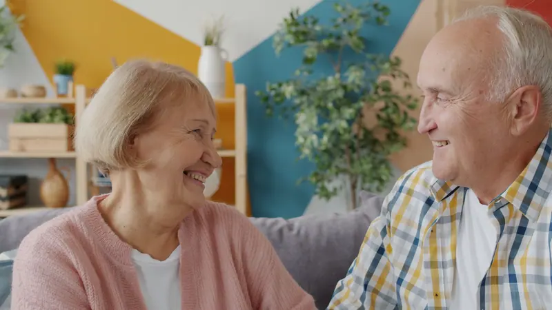 A resident and her friend laughing together on the sofa in the sunroom