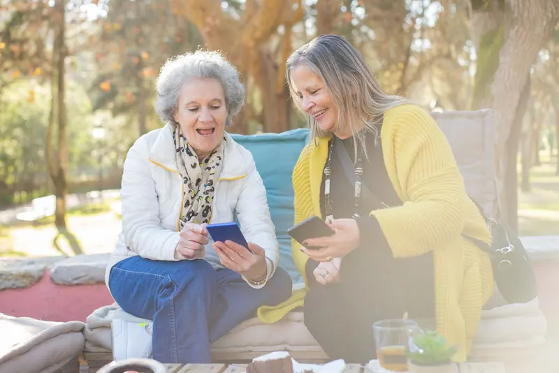 Two friends laughing over their phones on a sunny bench