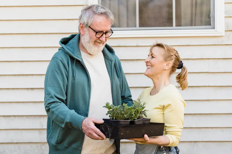 A couple carrying seedlings across the yard to their garden plot