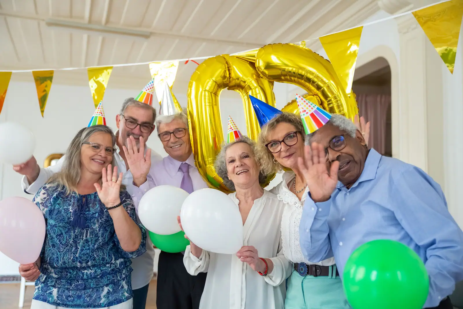 Residents in party hats waving and laughing under balloons and bunting at a clubhouse birthday party