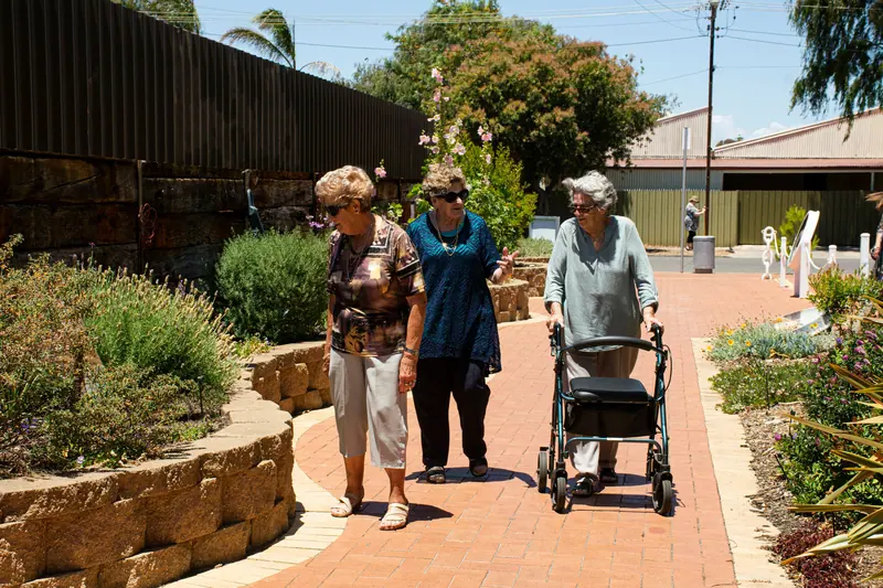 Three senior women strolling and chatting in a sunny garden setting, enjoying outdoor interaction.