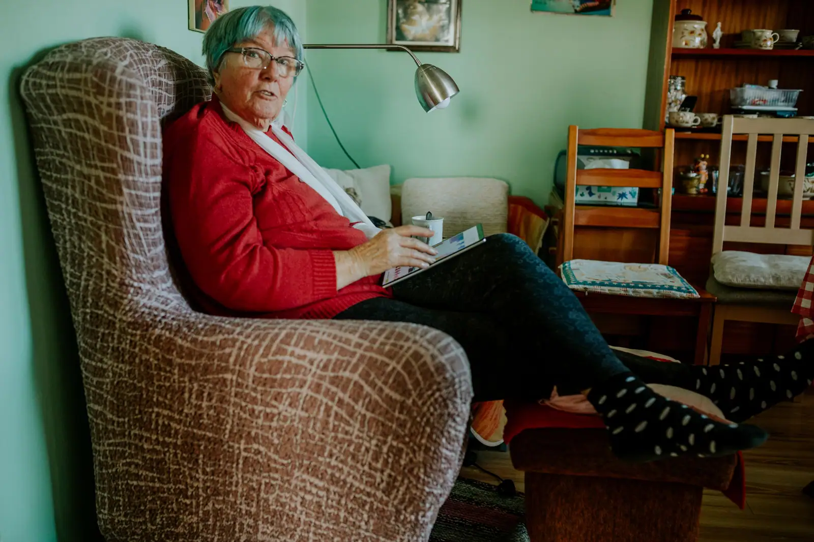 A resident in a red cardigan settled in her armchair with a book in her room
