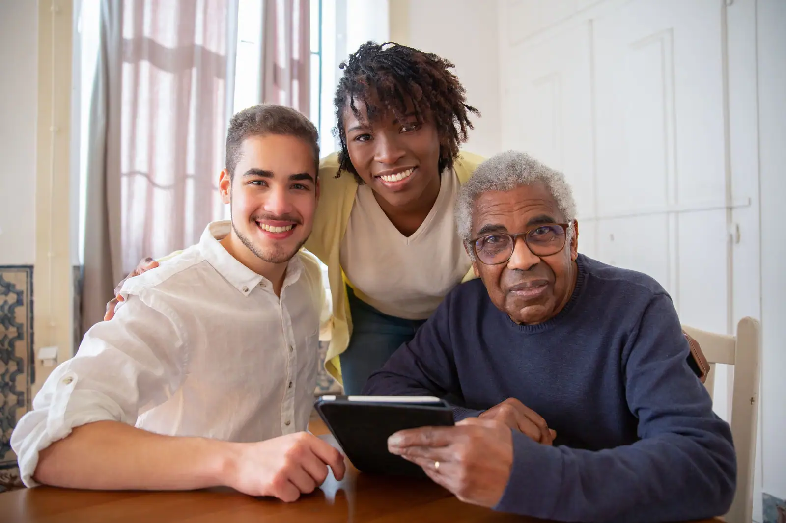 A resident smiling at a tablet with his grandson and a caregiver leaning in beside him