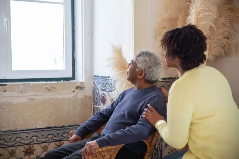 An aide in a yellow cardigan sitting with a resident by the window