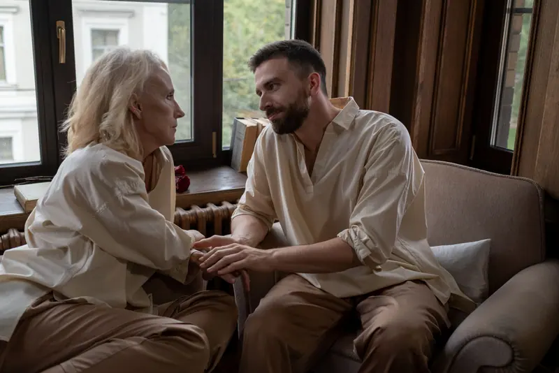 A grandson holding his grandmother's hands during a visit by the window